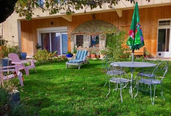 A grassy garden area features a round table with metal chairs, a green umbrella, and a chaise lounge. Two pastel-colored chairs are positioned nearby. The backdrop includes a wooden facade with large glass doors, allowing natural light to enter.