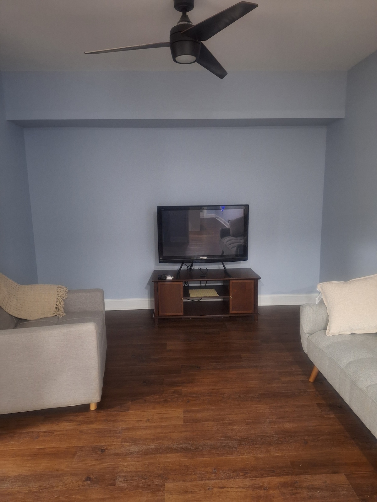 A light blue living room is presented, featuring two soft couches positioned across from a modest wooden entertainment unit. A television sits on the unit, while a ceiling fan is visible above. Natural light enhances the wood flooring.