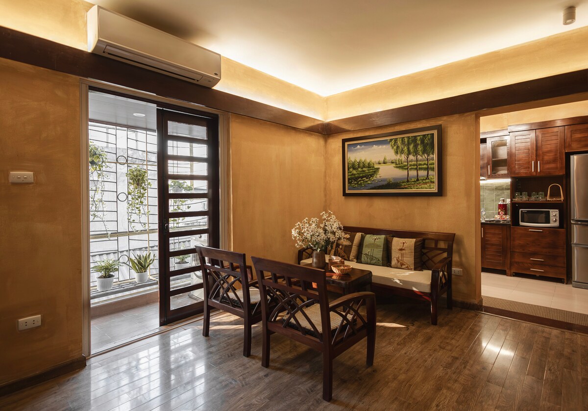 A welcoming living space with wooden seating positioned near a glass door leading to a balcony. Natural light flows into the room, complemented by warm-toned walls. A colorful painting of trees adorns the wall, enhancing the serene ambiance of this peaceful apartment.