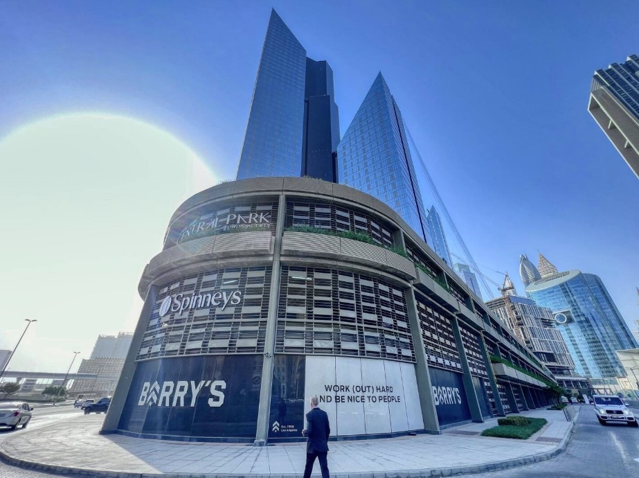 A modern building stands tall against a clear blue sky. The structure features large glass windows and an inviting entrance. A pedestrian is seen walking in front of the building, which houses retail spaces including a supermarket and a fitness center.