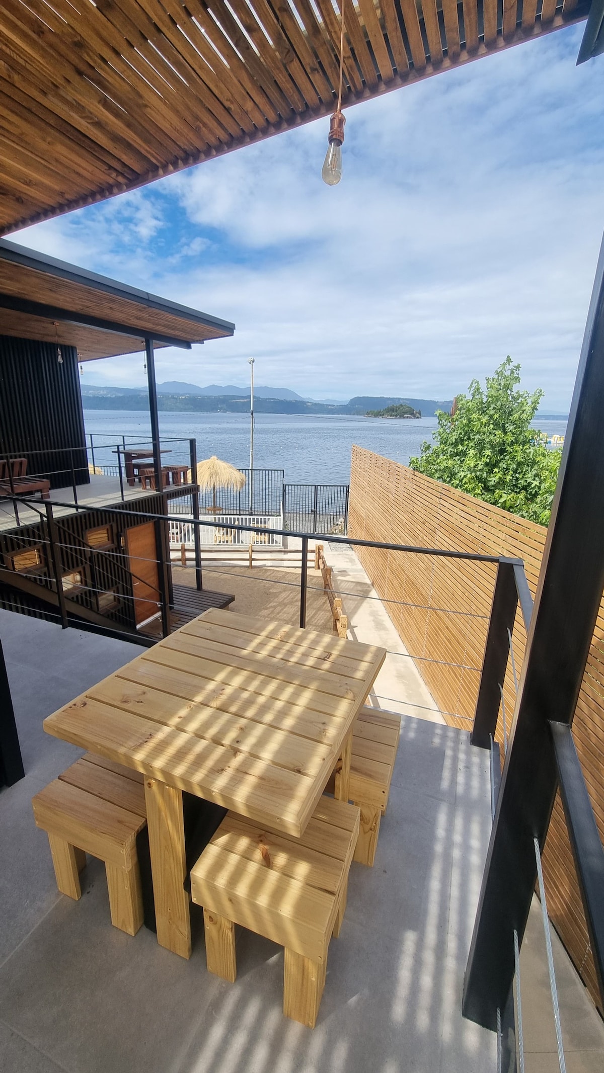 An outdoor dining area is depicted with a light wooden table and matching stools, positioned on a balcony. Scenic views of the lake and distant mountains are visible in the background, complemented by a clear sky and natural light filtering through the overhead slats.