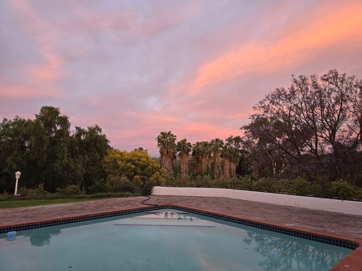 A serene view of the swimming pool reflects the colorful sky at dusk. Lush greenery, including tall palm trees and blooming flora, enhances the tranquil setting against the backdrop of a softly lit horizon.