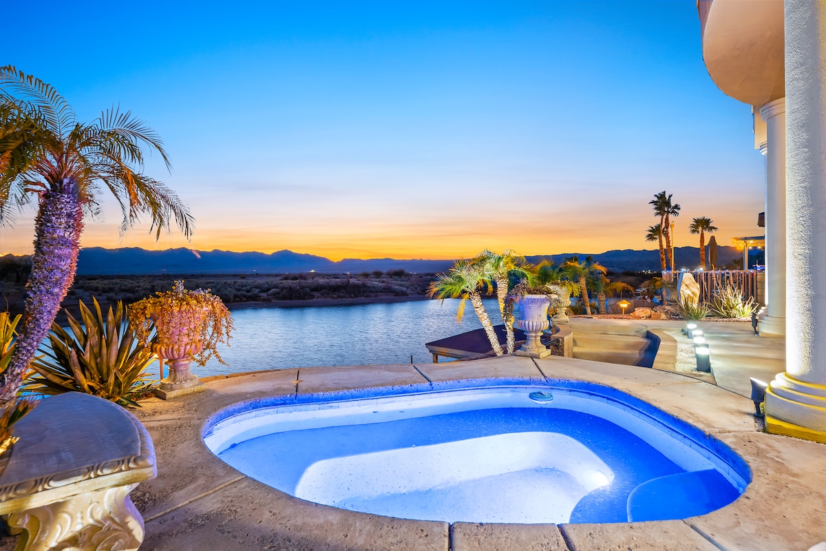 A hot tub is situated near the water's edge, surrounded by tropical plants and palm trees. The scene captures a sunset, with vibrant colors reflected in the sky above the river. Comfortable seating areas are visible in the background, enhancing the outdoor relaxation space.