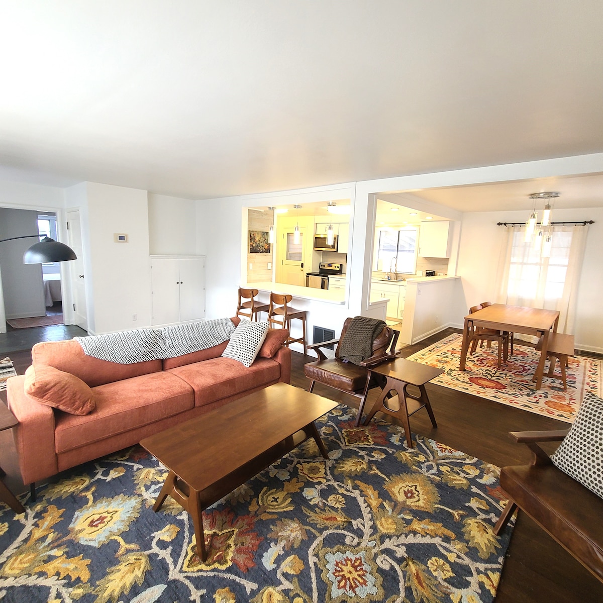 A bright living area features a cozy orange sofa and a wooden coffee table, surrounded by patterned rugs. Light enters through large windows, illuminating the open layout that connects to a dining space with a table and chairs in the background.