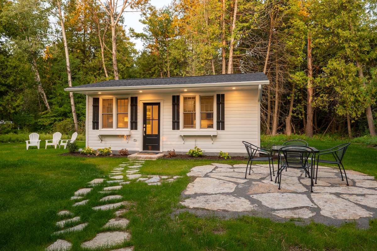 The exterior of a charming cabin is featured, set within a lush green landscape. A stone path leads to the entrance, flanked by well-maintained grass. Outdoor seating consists of a round table surrounded by black chairs, and two white chairs are positioned nearby.