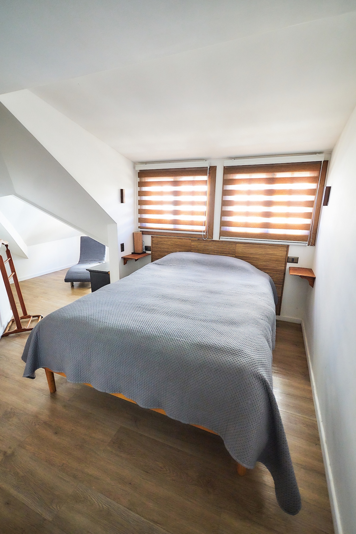 A cozy bedroom features a spacious bed covered with a light gray blanket. Two large windows with wooden blinds allow natural light to filter in. A small chair is positioned in a corner, and a modern wooden headboard enhances the room's simplicity.