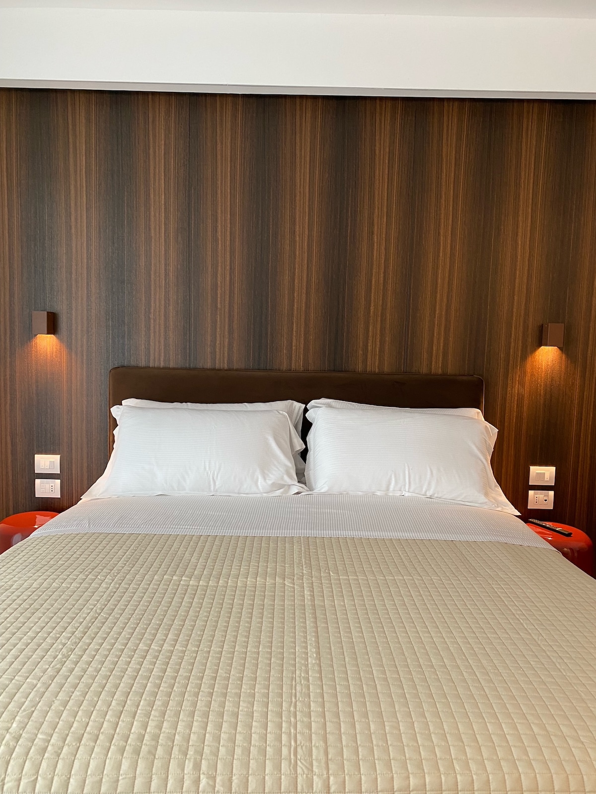 A cozy sleeping area features a spacious bed with light-colored bedding, complemented by two white pillows. The brown wood paneling creates a warm backdrop, with subtle lighting fixtures flanking the bed, adding to the serene ambiance.