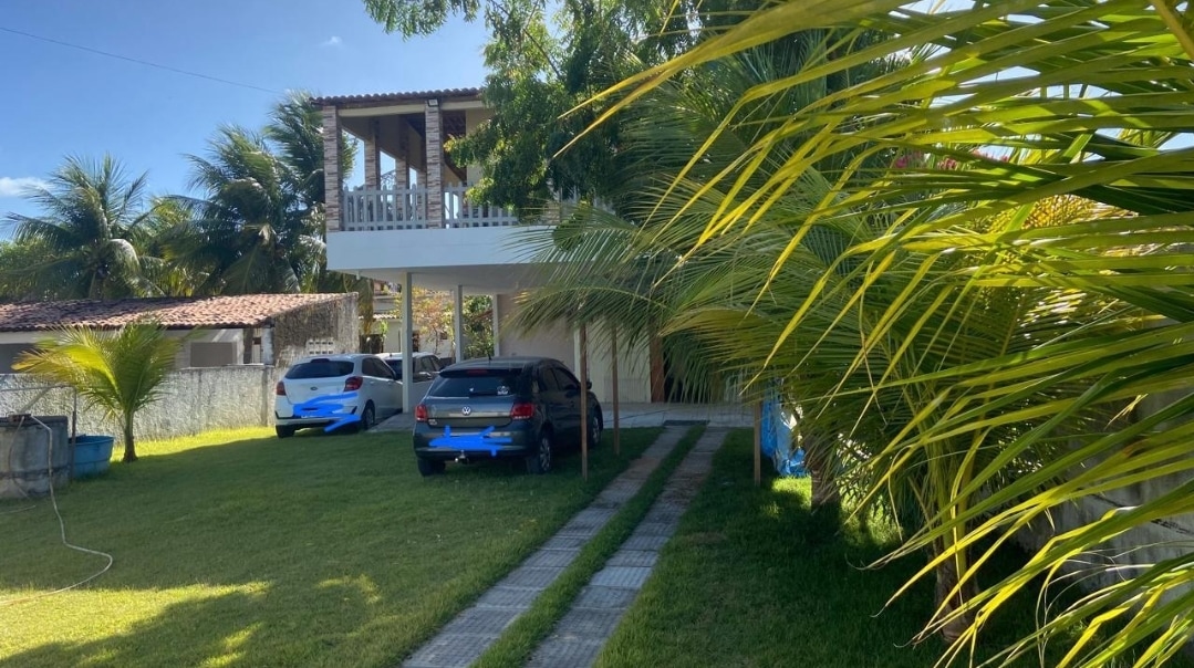 A spacious exterior view features a two-story structure surrounded by lush green palm trees. A driveway leads to the house, with two parked cars visible. A balcony on the upper level offers an outdoor view, set against a clear blue sky.