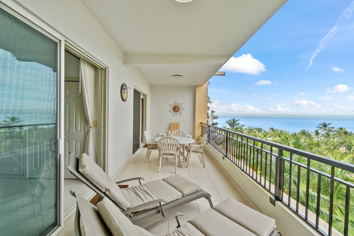A spacious balcony features a round dining table surrounded by four chairs. Two lounge chairs are positioned for relaxation, with views of lush greenery and the ocean in the background. Large sliding doors provide access to the interior, allowing natural light to fill the space.
