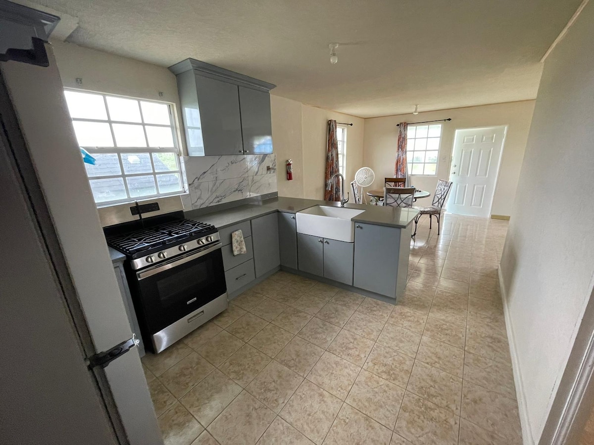 An open-concept kitchen is visible, featuring a modern gas stove, sleek cabinetry, and a spacious countertop. Large windows allow natural light to fill the area, illuminating the dining set and inviting seating in the background. Tiled flooring enhances the bright atmosphere.