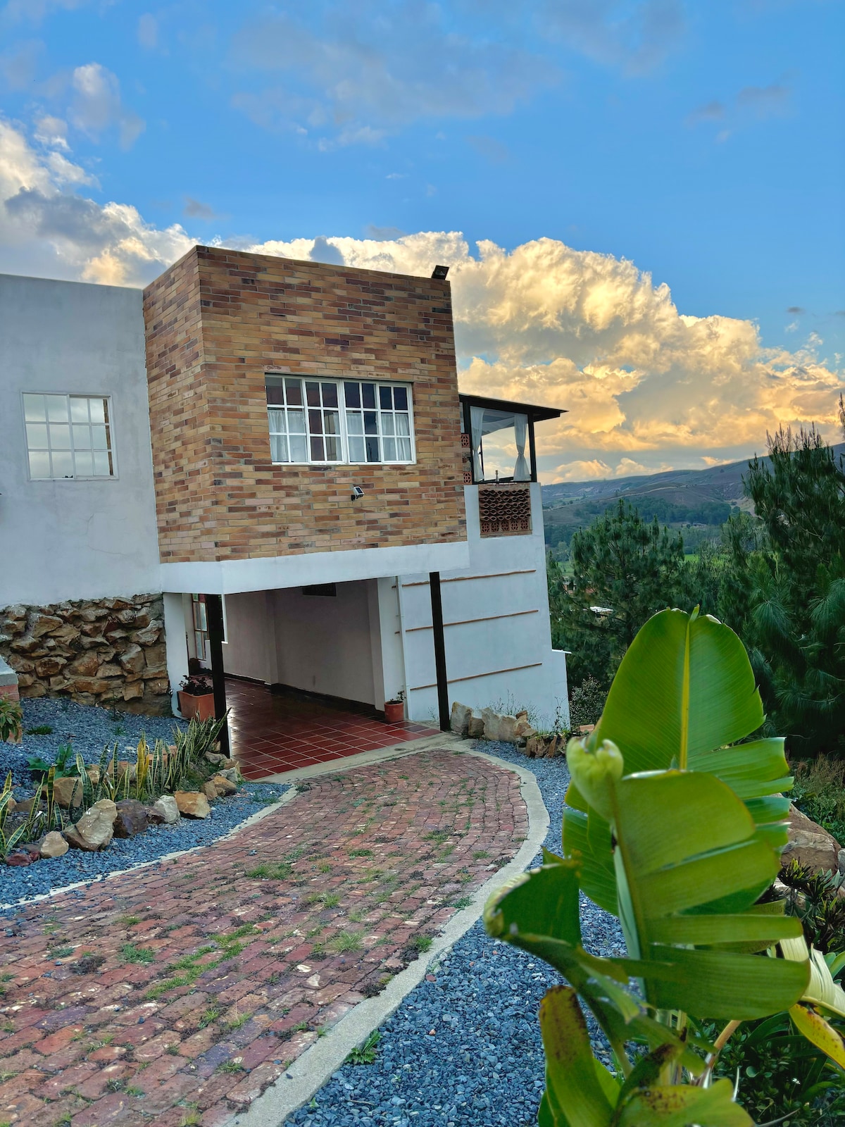 A modern house is presented against a backdrop of mountains and a colorful sky. The structure features a mix of brick and smooth surfaces, with large windows allowing natural light inside. A winding pathway leads to the entrance, bordered by lush greenery and pebbled landscaping.