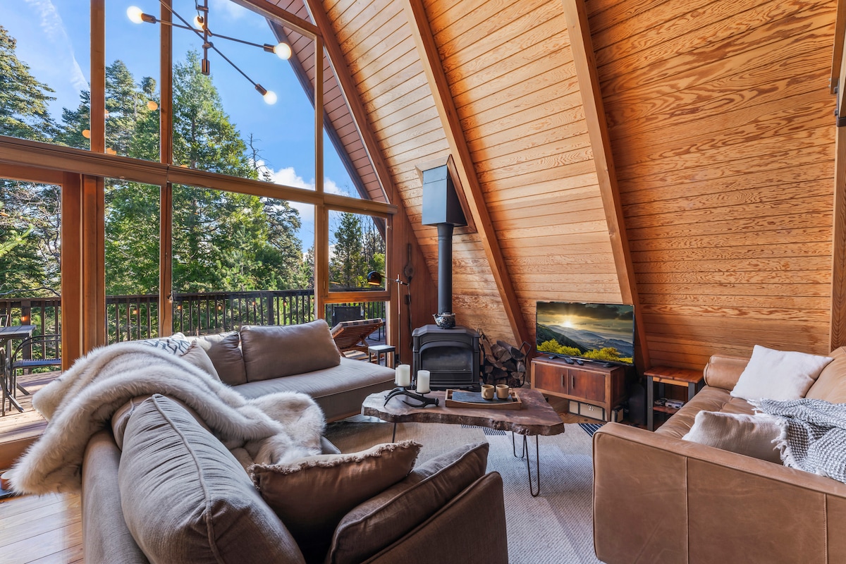 The living space features high vaulted ceilings and large windows that invite ample natural light, offering views of the surrounding trees. Cozy seating arrangements include a plush sectional and a leather couch. A wooden coffee table centers the area, while a wood stove provides warmth.
