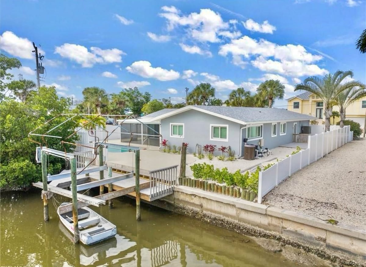 A serene canal view is presented, showcasing a boat dock with a small boat anchored nearby. The beach house, painted in soft grey, features multiple windows and is surrounded by lush greenery and well-maintained landscaping, providing a tranquil waterfront setting.