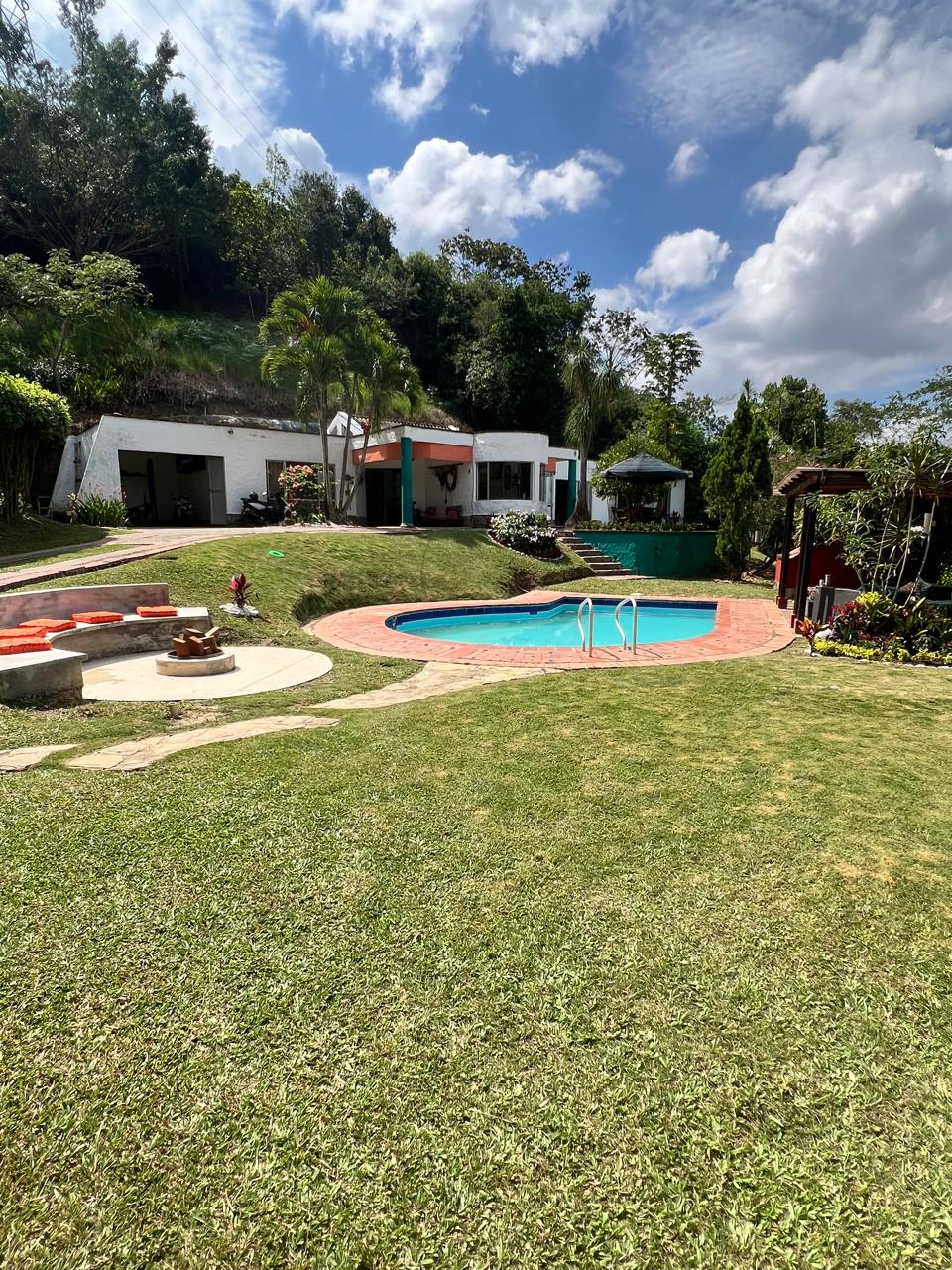 A lush green lawn leads to a vibrant swimming pool surrounded by terracotta tiles. Lounge chairs are positioned nearby, and a fire pit is visible in the foreground, framed by a stone seating area. The backdrop features charming landscaped gardens and inviting outdoor spaces.