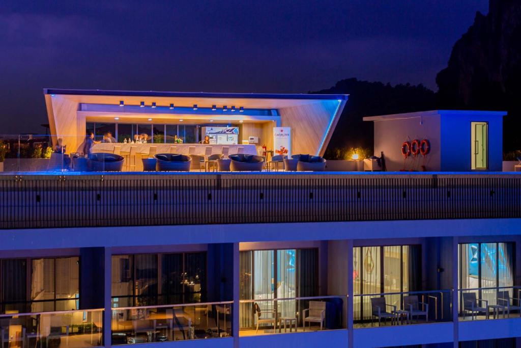 An illuminated rooftop bar is showcased with modern seating and a sleek design. The space is framed by a decorated railing and offers a serene ambiance against a darkening sky. The building below features glass balconies, providing glimpses of the welcoming indoor spaces.