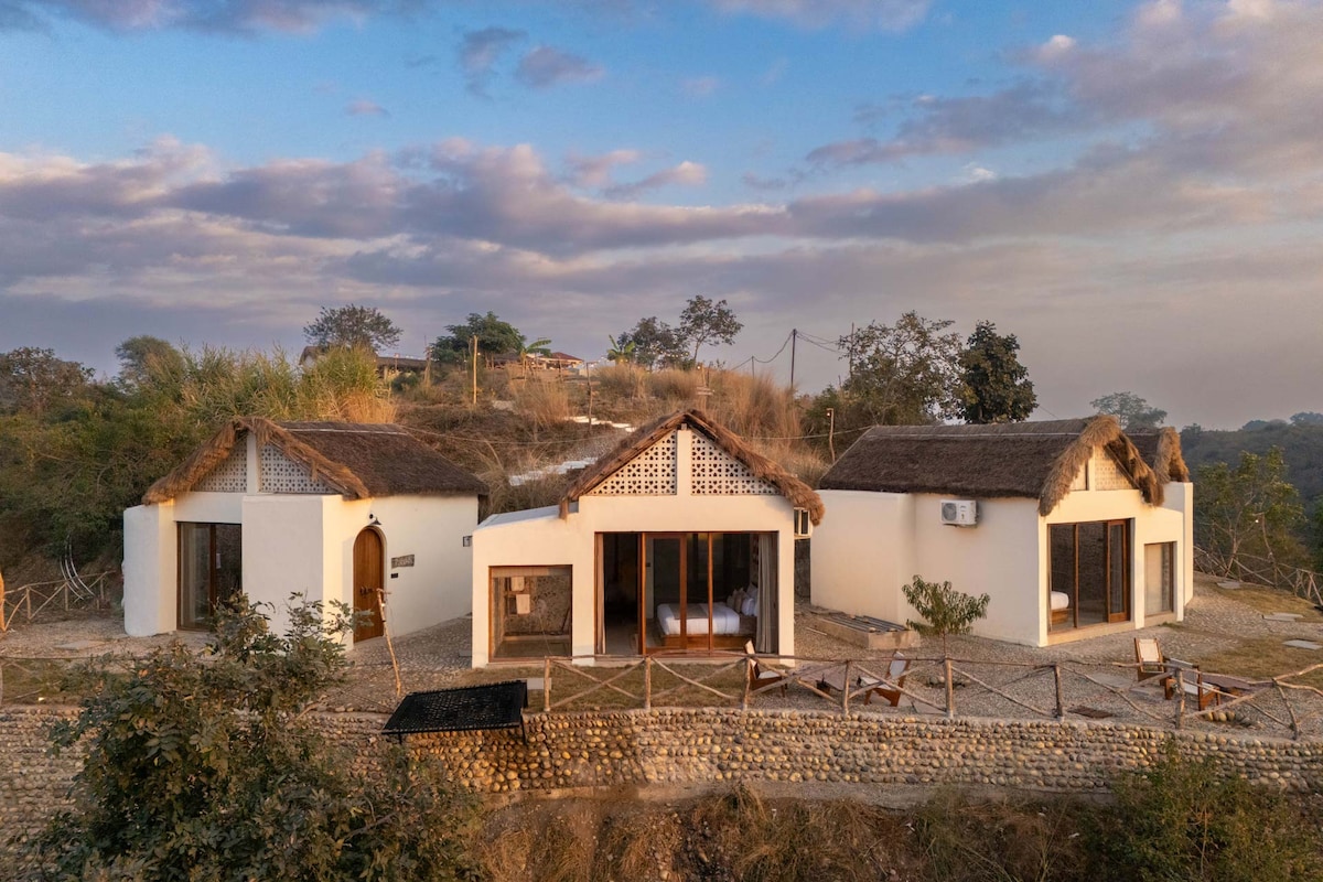 Two individual cottages are nestled within natural surroundings, featuring thatched roofs and large glass doors that provide scenic views. A gravel path leads to the cottages, while lounge chairs are arranged outside, inviting moments of relaxation in the serene landscape.