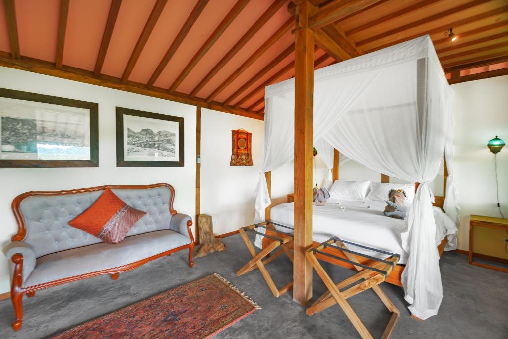 A spacious bedroom is furnished with a large canopied bed draped in sheer fabric. A gray sofa with an accent pillow is positioned nearby. Natural light enters through the window, highlighting wooden beams and a decorative rug on the floor.