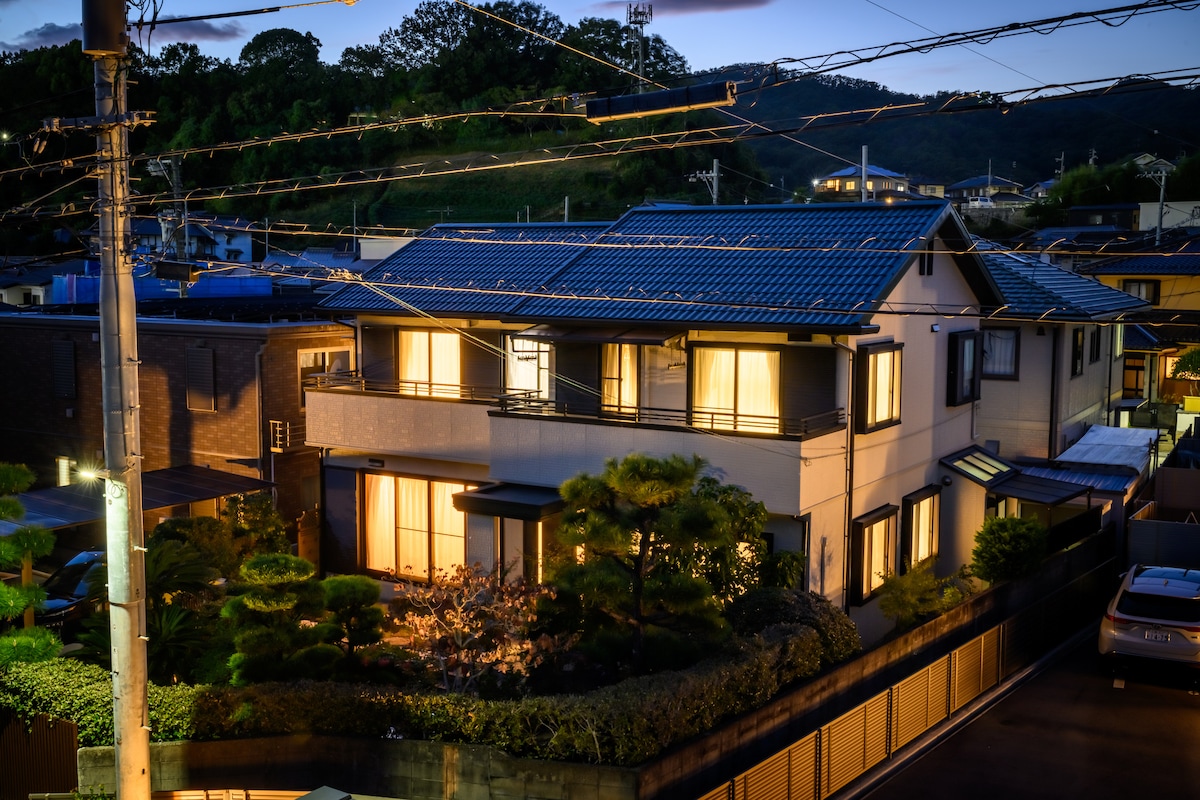 The exterior of a two-story home is illuminated at dusk, showcasing large windows that reflect warm light. Solar panels are visible on the roof. A neatly maintained garden with trees and shrubs surrounds the property, enhancing the serene ambiance.