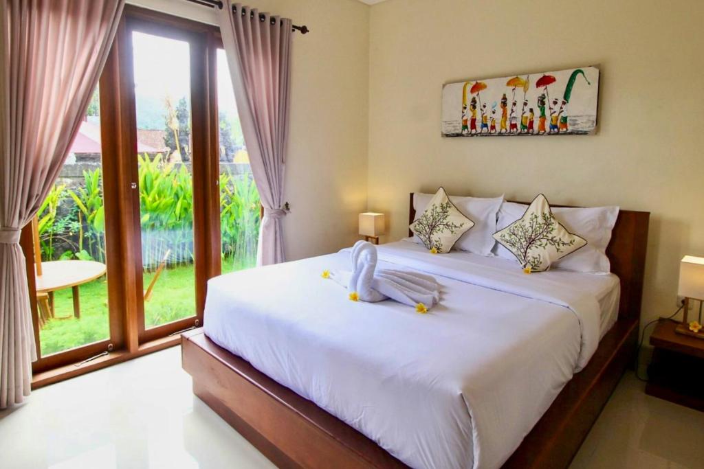 A spacious double room is featured, furnished with a large bed adorned with decorative pillows. Natural light enters through large windows, revealing a view of the garden outside. A small table and chairs are positioned next to the windows, creating a cozy atmosphere.