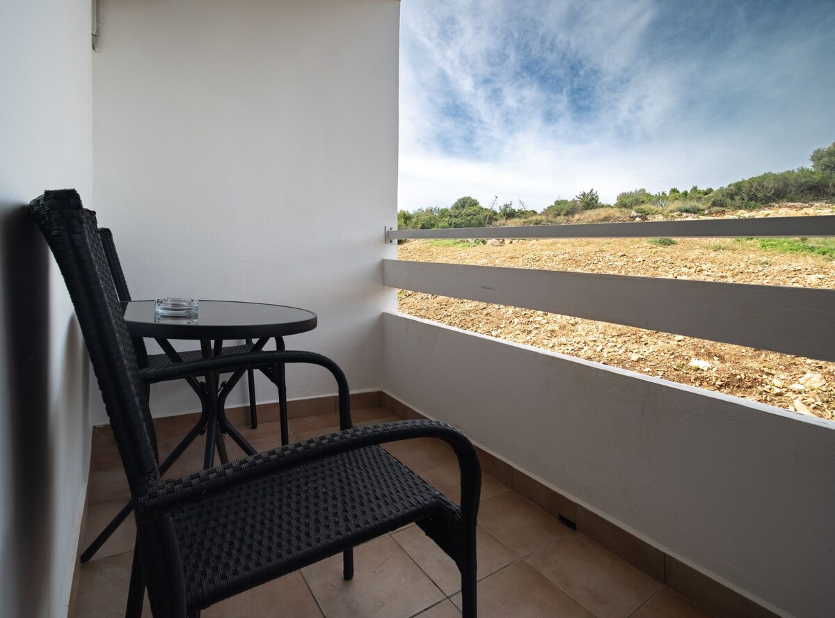 A balcony area features two black wicker chairs positioned around a small table, providing a space for relaxation. The white railing offers a view of a natural landscape, which includes greenery and a gravel path under a clear sky.