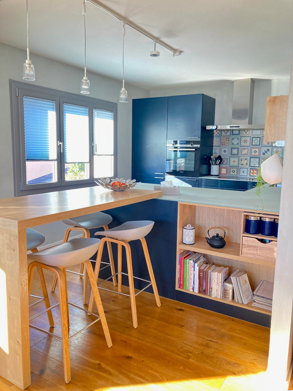A modern kitchen area showcases a spacious bar with three wooden stools. The cabinetry features a blend of dark and light tones, complemented by patterned tiles. Large windows illuminate the space, offering views of the outside while providing natural light.