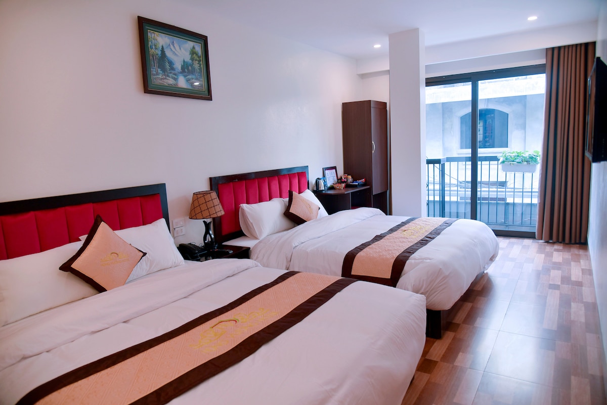 The room features two spacious beds with elegant headboards and embroidered bedding. A sliding glass door provides access to a private balcony, allowing natural light to fill the space. A wooden wardrobe and a small dressing table are positioned nearby.