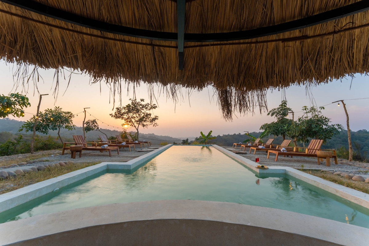 An infinity pool stretches toward the horizon, reflecting the soft hues of sunset. Wooden lounge chairs with plush cushions are situated around the edge, inviting relaxation. Lush greenery flanks the pool area, enhancing the serene outdoor atmosphere.