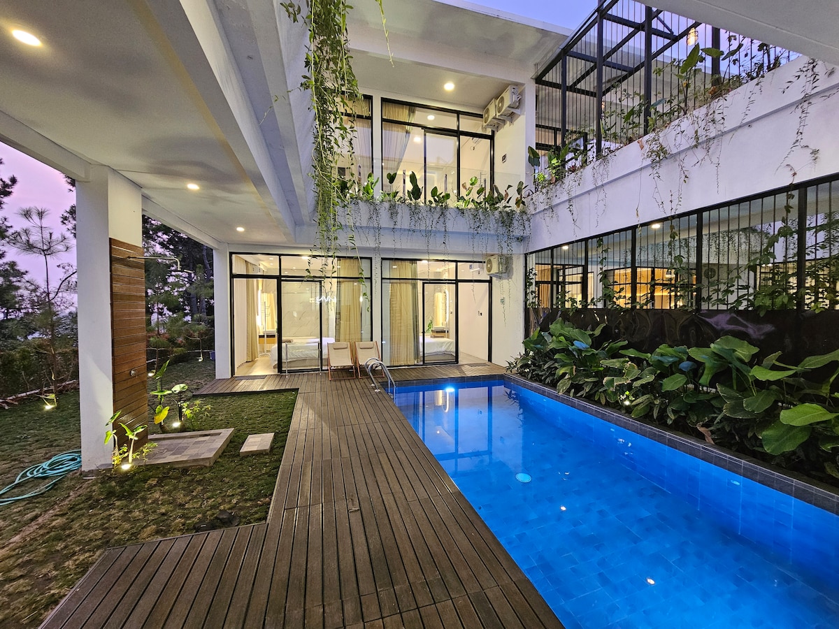 A serene outdoor area features a private pool with blue water, surrounded by wooden decking and lush greenery. Large glass doors lead into the villa, inviting natural light into the interior spaces. Vines hang gracefully from the upper balcony, adding to the tranquil setting.
