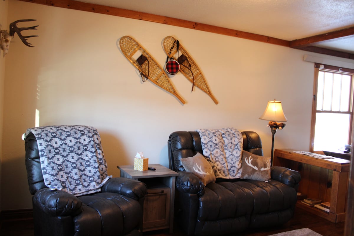 A cozy living area features two black reclining chairs draped with patterned blankets. Above them, vintage snowshoes and a decorative red plaid ball add character to the wall. A small wooden table is positioned between the chairs, completing the inviting space.