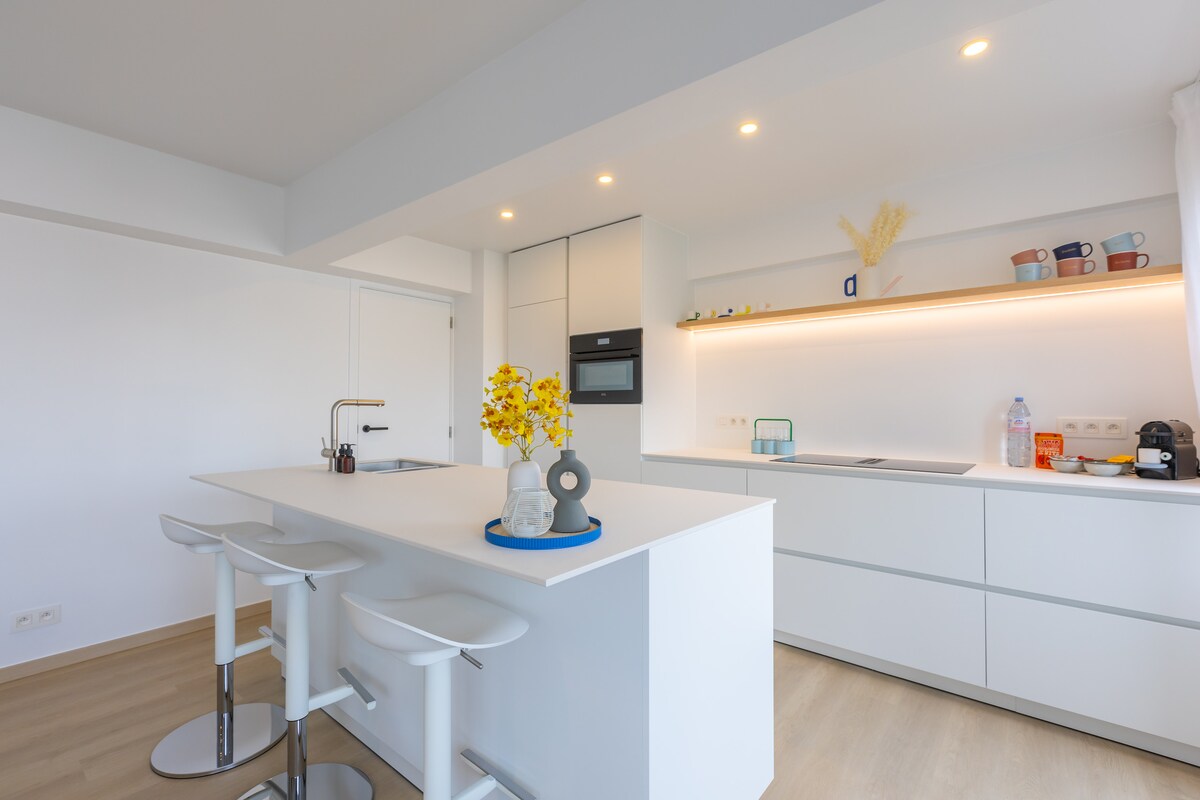 A modern kitchen features sleek white cabinetry and a spacious central island with three high-backed stools. Soft lighting highlights decorative dishware on the shelves, while a sleek oven and minimalistic appliances complete the inviting atmosphere.
