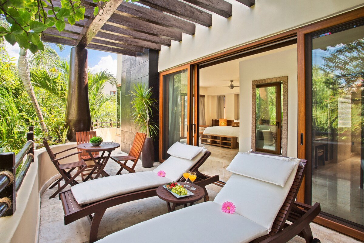 A private terrace is highlighted by two wooden lounge chairs and a small table, surrounded by lush greenery. Sunlight filters through the overhead beams, enhancing the connection between indoor and outdoor spaces. A refreshing drink and tropical flowers are arranged on the table.