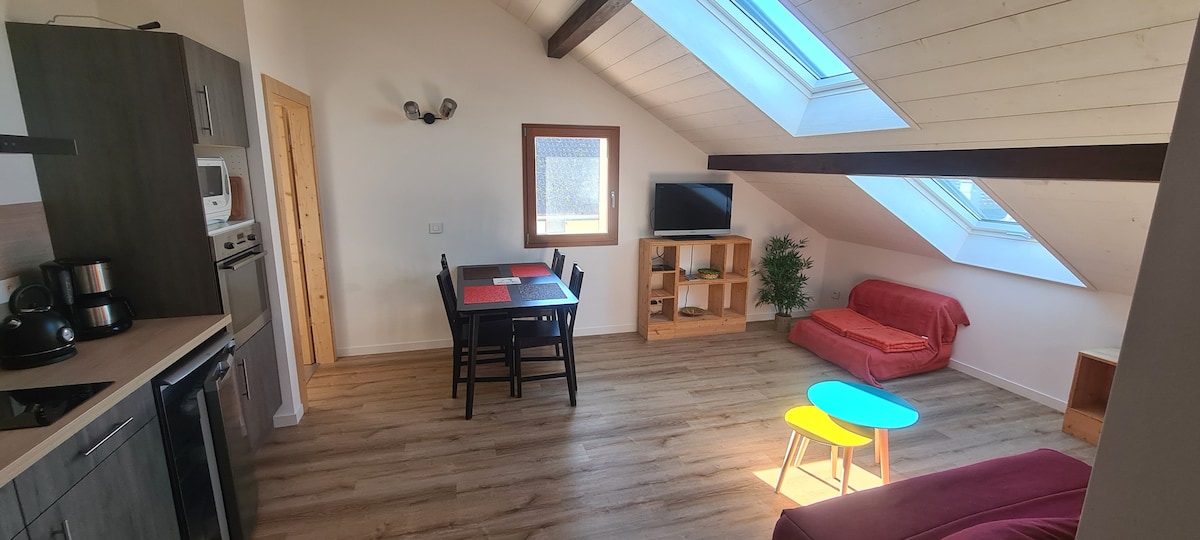 The living area features sloped ceilings with skylights allowing ample natural light. A dining table with four chairs and a modern kitchen with appliances are visible. A small sofa bed provides additional seating. A TV is positioned on a shelf near a decorative plant.