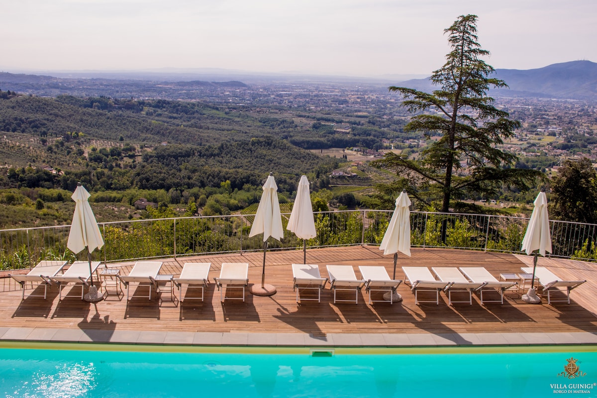 A serene outdoor pool area is visible, featuring several lounge chairs arranged on a wooden deck. White umbrellas provide shade, and a panoramic view of the lush valley and distant hills is captured in the background.