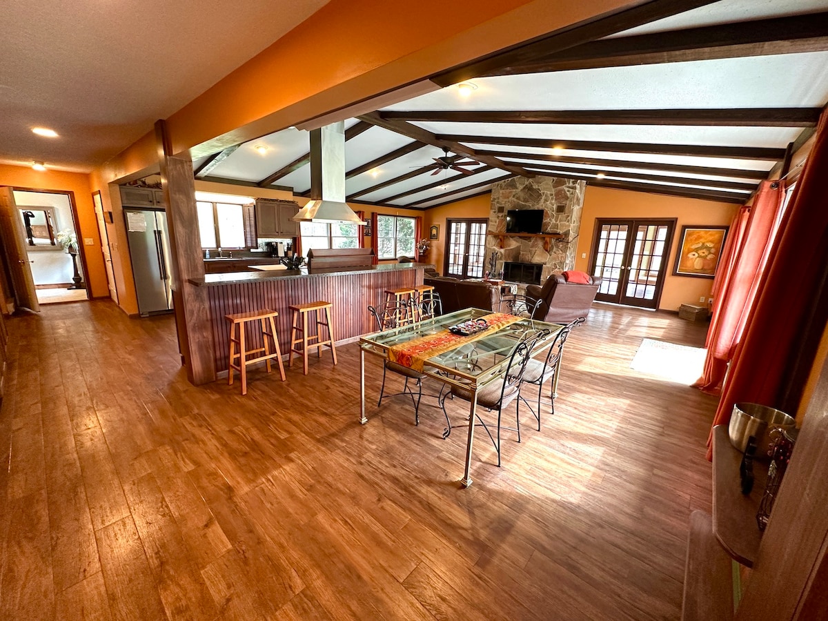 An open and airy living space is depicted, featuring exposed wooden beams across the ceiling. A glass dining table is set with iron chairs, while a cozy seating area with a stone fireplace and large windows invites natural light.