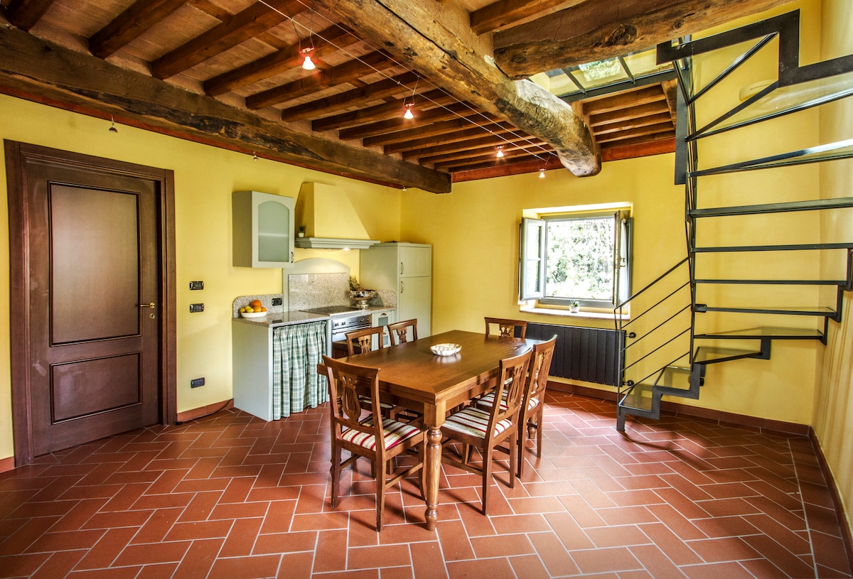 A dining area features a wooden table surrounded by chairs, with natural light entering through a window. An open kitchen with modern appliances and a stone countertop is visible, complemented by rustic wooden beams above. A contemporary spiral staircase leads to the upper level.