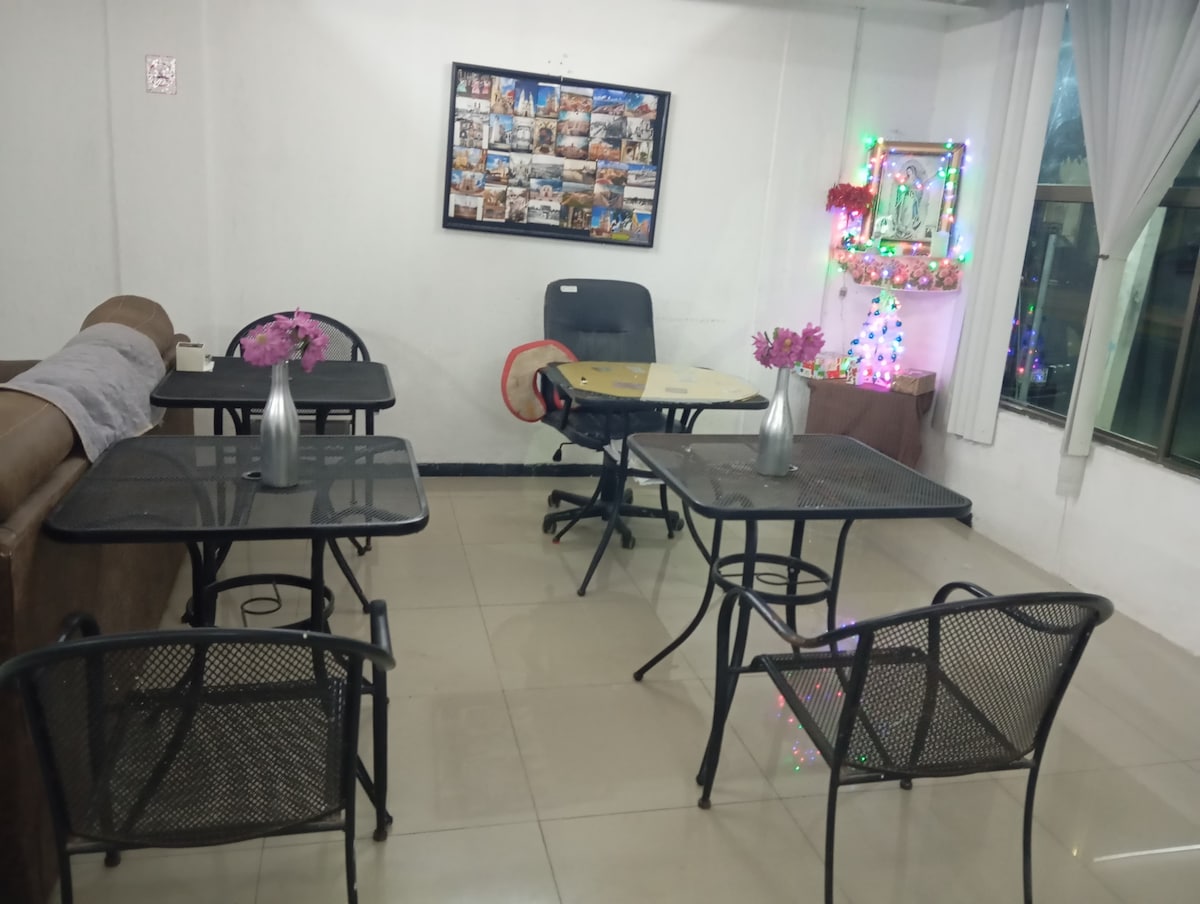 A dining area is configured with four black metal tables and chairs arranged neatly, featuring vases with flowers. A wall is adorned with a photo collage, while a small decorative tree with lights adds a festive touch to the space.