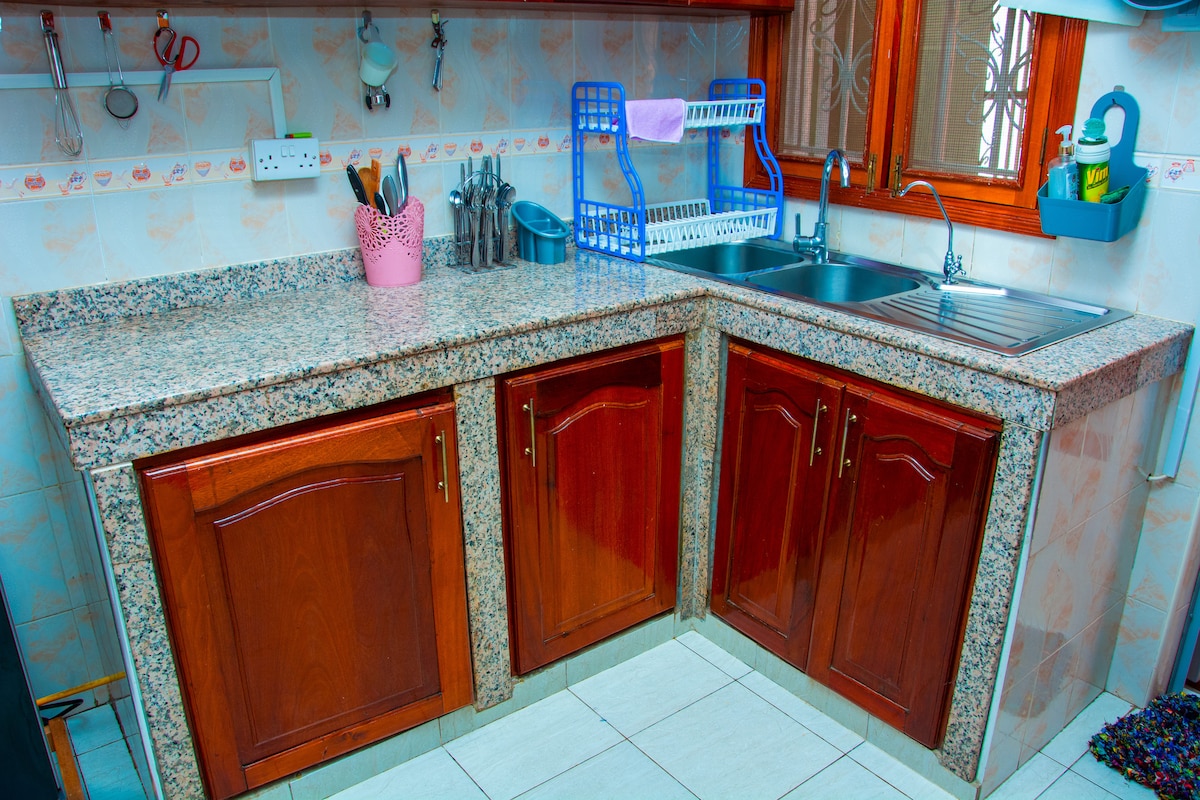 A well-organized kitchen area features a granite countertop with wooden cabinetry beneath. Stainless steel utensils and a sink are visible, alongside a dish rack holding dishes. Natural light enters through a window adorned with a sheer curtain, enhancing the workspace.