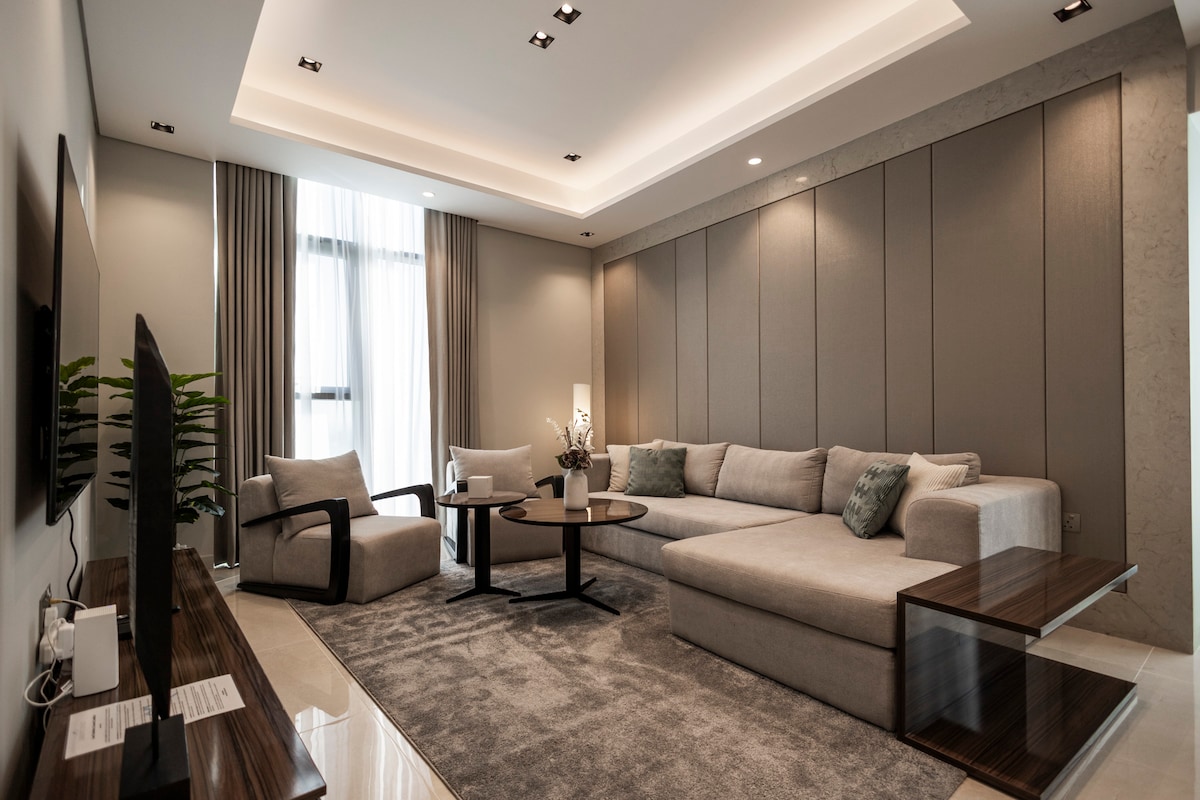 A spacious living area is designed with neutral tones, featuring a large sectional sofa and an elegant coffee table. Natural light floods the space through large windows, complemented by soft curtains. A flat-screen TV is mounted on the wall, adding to the modern design.