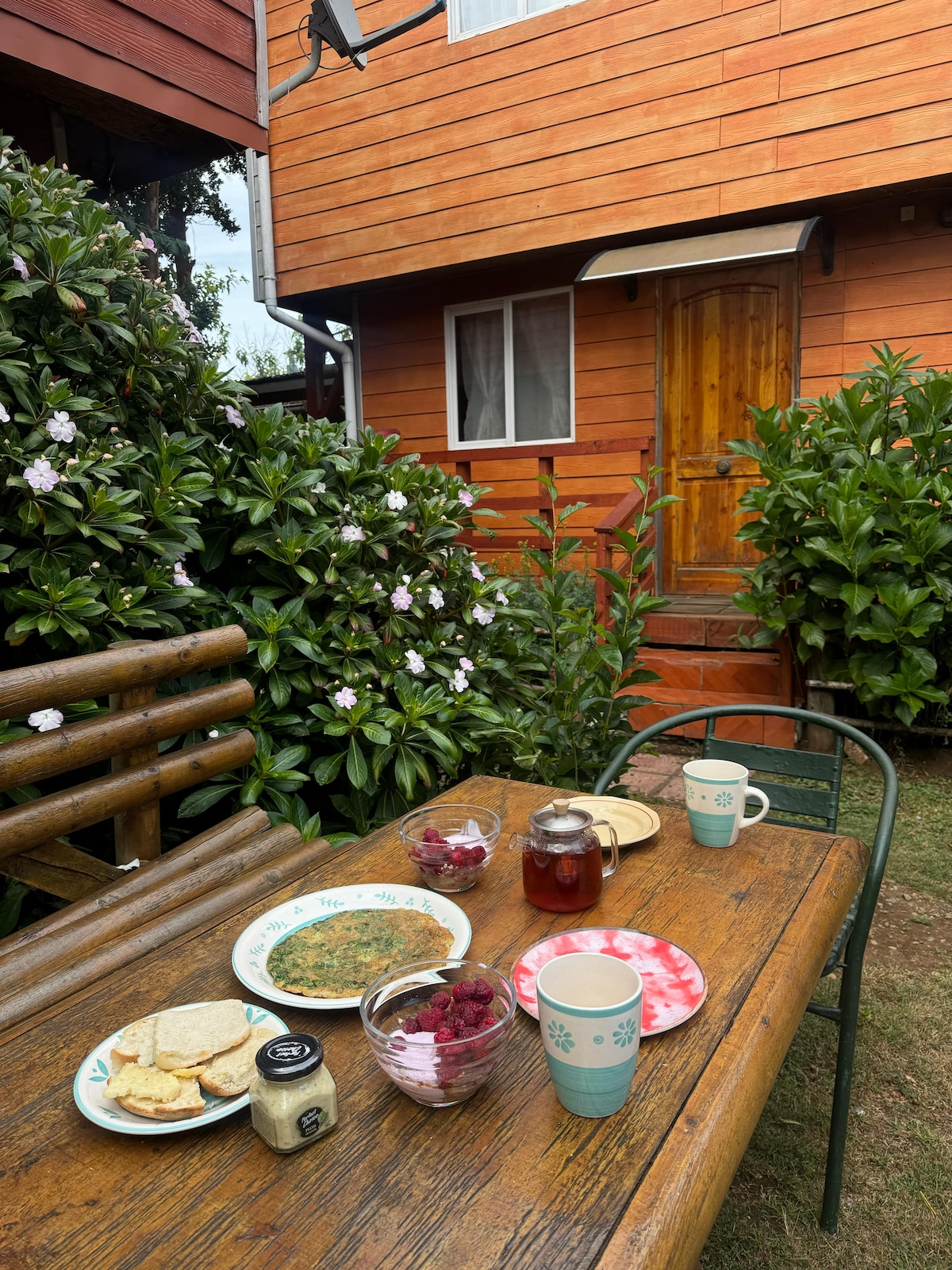 A rustic wooden table is set for breakfast outside, featuring plates with toast and a colorful bowl of fresh raspberries. Cups and a jar of jam are arranged among lush greenery and flowering shrubs, with a wooden cabin visible in the background.