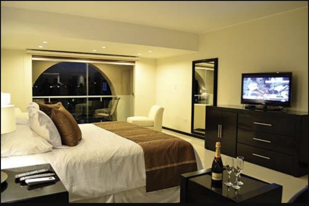 A spacious bedroom features a king sized bed dressed in neutral linens, complemented by decorative pillows. A large window offers ample natural light, while a wall-mounted television and a dark wood dresser add functionality. A small seating area is set beside the window.