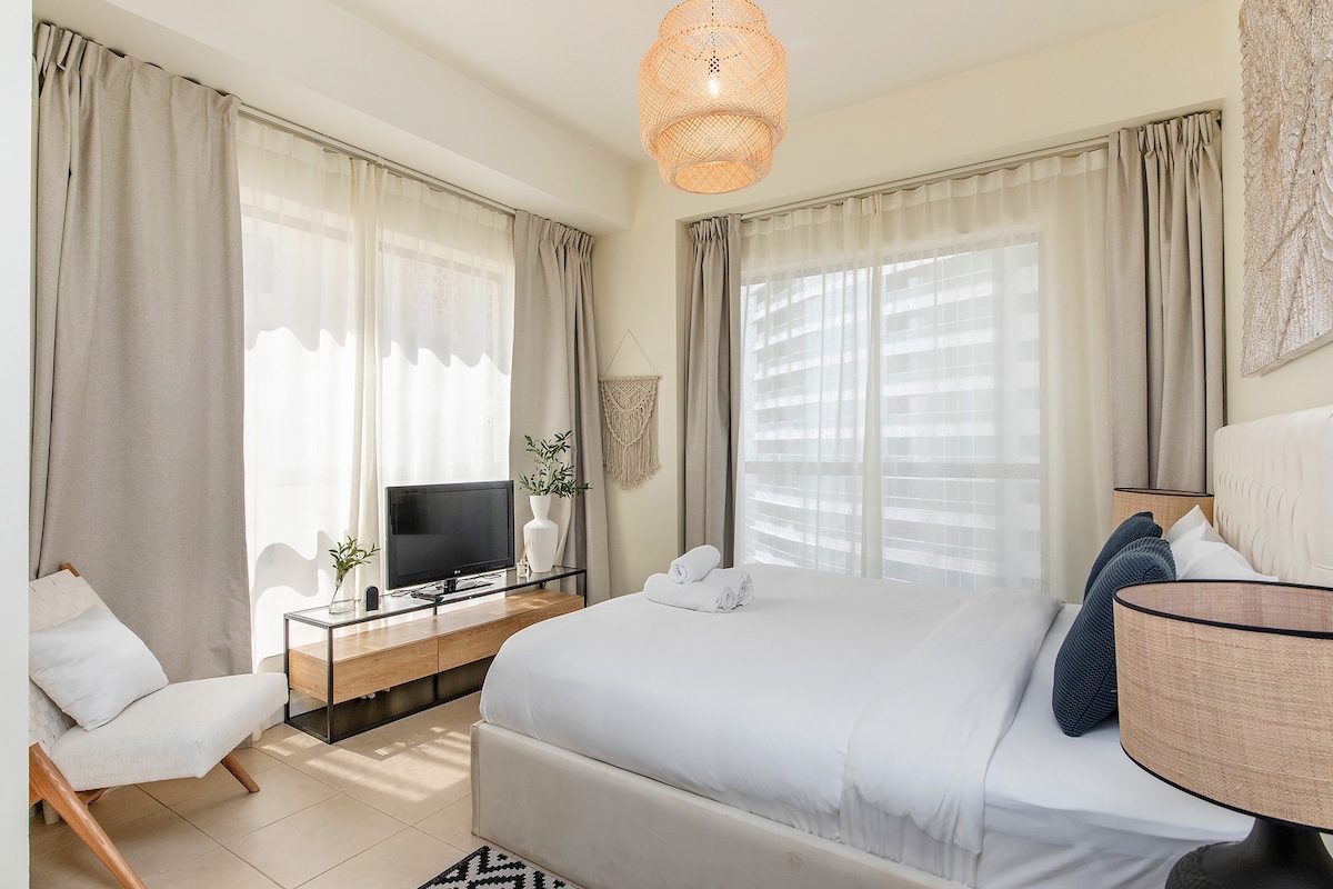 A well-appointed bedroom features a spacious bed adorned with white bedding, complemented by a minimalistic decorative wall piece. Light floods in through large windows, draped with sheer curtains. A flat-screen TV is positioned on a contemporary glass table, while a cozy armchair adds comfort to the inviting space.