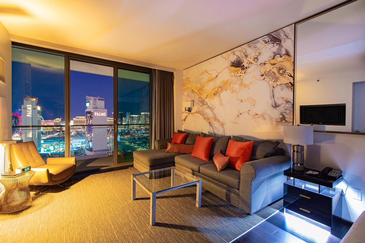 An open-concept living area features a comfortable gray sofa with bright orange accent pillows, a glass coffee table, and a sleek side chair. Floor-to-ceiling windows reveal views of the vibrant Las Vegas Strip at dusk, complemented by a contemporary marble wall design.