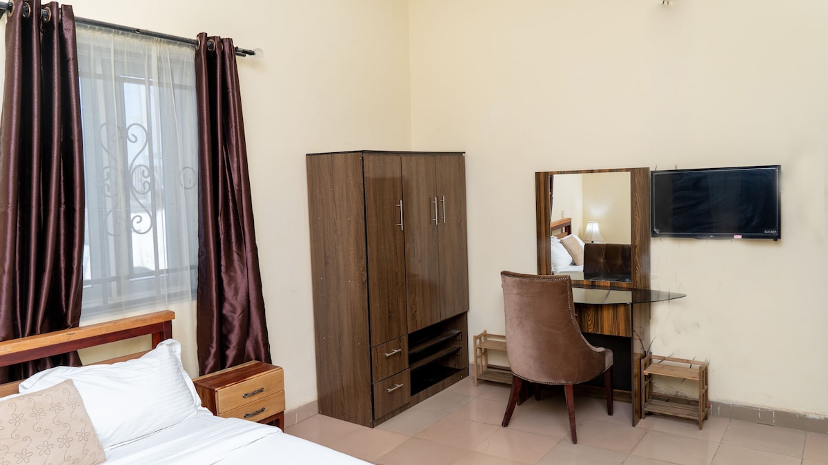 A cozy bedroom features a spacious bed with white linens, complemented by wooden nightstands. A large wardrobe stands against the wall, next to a mirrored dressing table with a comfortable chair. Natural light filters through the window, adorned with dark curtains.