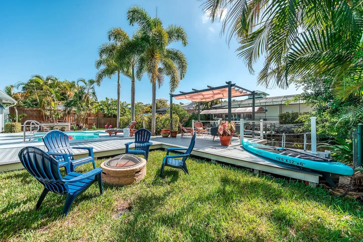 A deck area is complemented by several blue Adirondack chairs arranged around a circular fire pit. Lush greenery surrounds the space, with palm trees and decorative planters visible. A kayak rests beside the deck, while a shaded pergola provides a relaxing area for outdoor enjoyment.