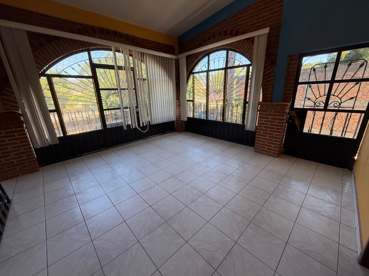 A spacious room features large arched windows with light filtering through sheer curtains. The tiled floor enhances the openness, and brick detailing adds warmth to the walls. Two sets of doors provide access to the outside, creating a harmonious connection with the surrounding environment.