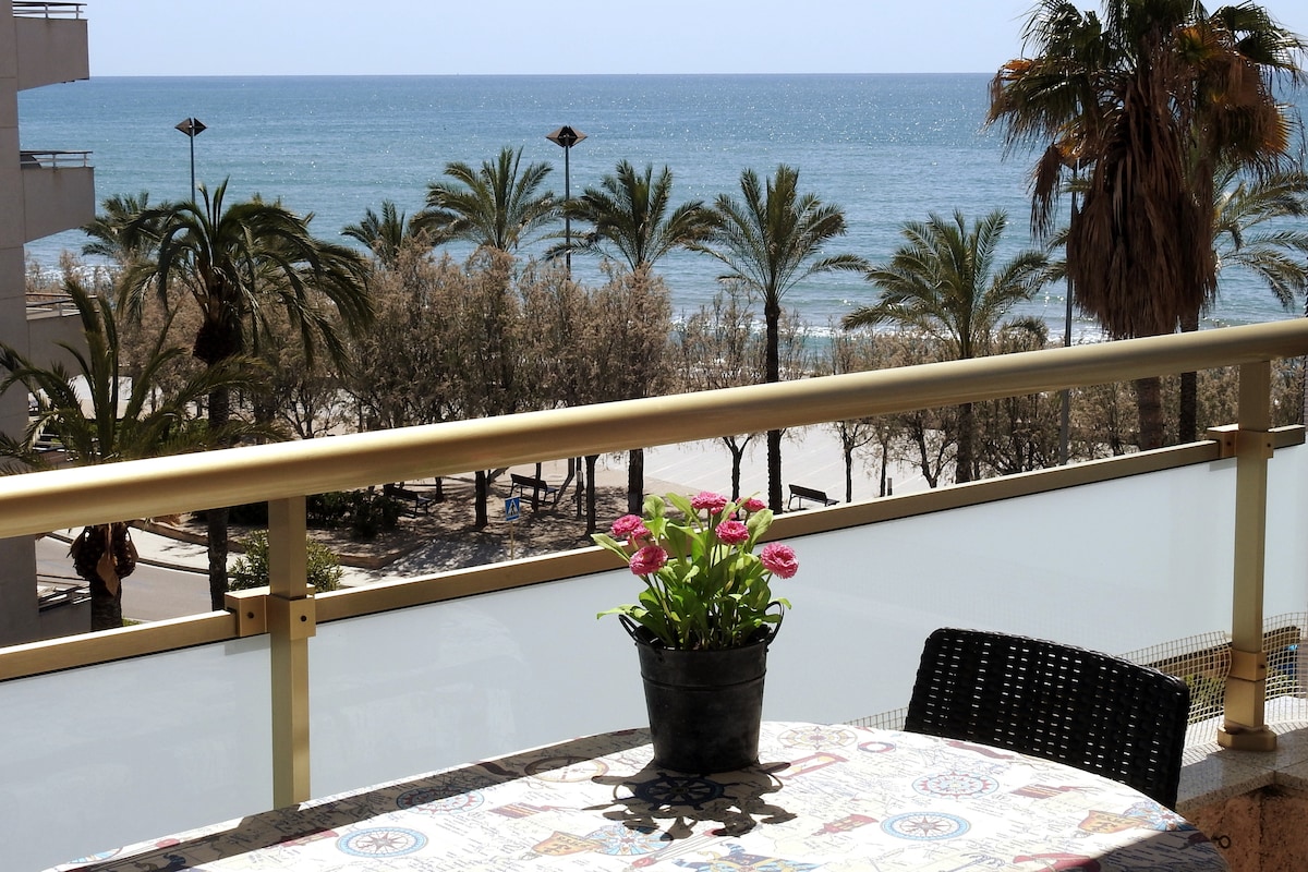 A spacious terrace features a table adorned with a potted plant, overlooking a scenic view of the beach and palm trees. The calming blue of the sea contrasts with the greenery, creating a refreshing atmosphere. The area is well-lit by natural sunlight.