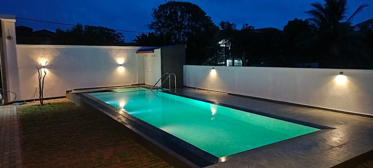 A tranquil swimming pool surrounded by a well-maintained outdoor area is illuminated at night. Soft lighting enhances the pool's water, casting a soothing turquoise glow. The surrounding walls provide privacy, and lush greenery is visible in the background.