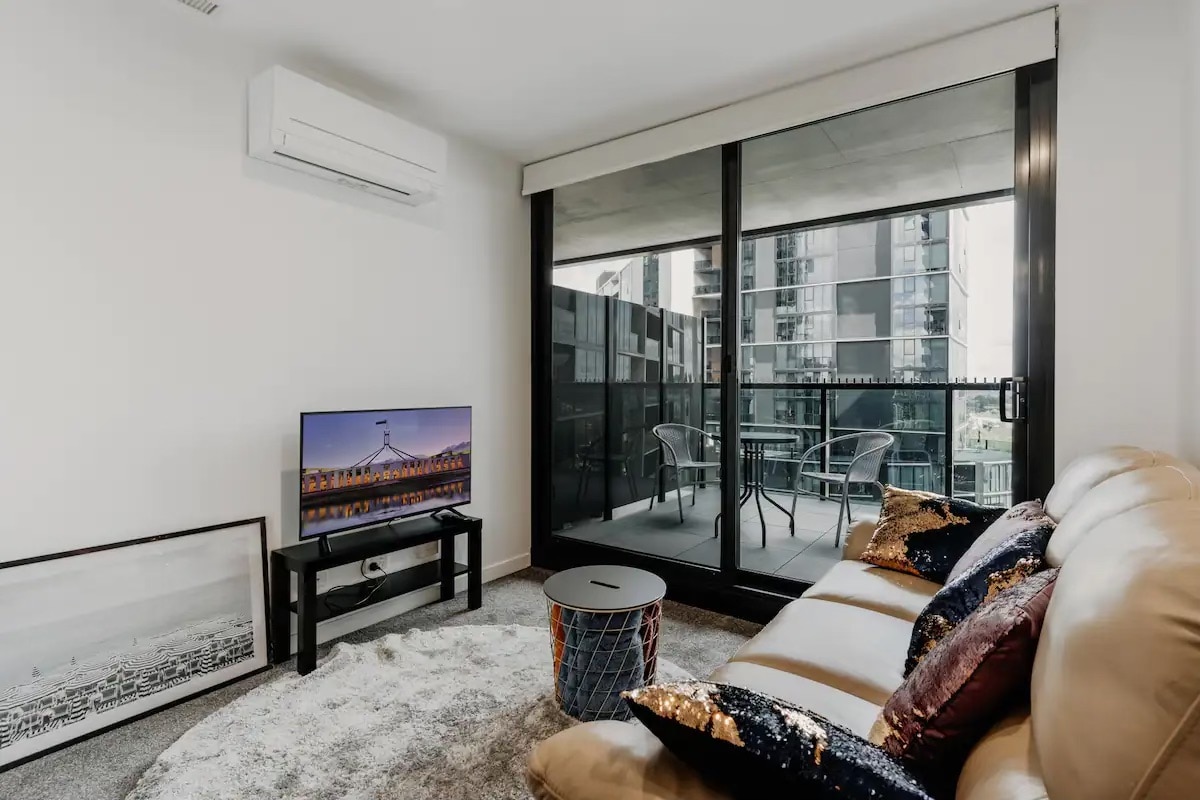 A comfortable living area features a plush sofa adorned with decorative pillows, alongside a contemporary coffee table. A television sits on a modern stand, and floor-to-ceiling glass doors lead to a private balcony, offering views of the surrounding buildings.