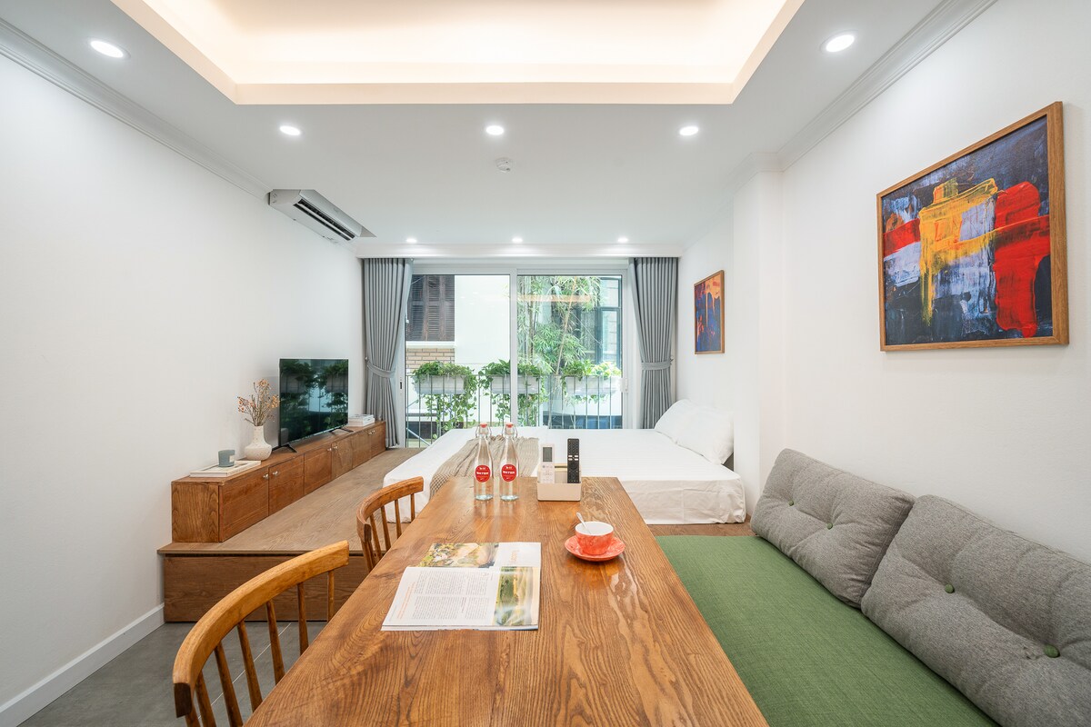 A bright living area is displayed, featuring a dining table with wooden chairs and a large piece of green seating. A Smart TV is positioned on a wooden console, while large windows invite natural light, offering views of greenery outdoors. Neutral tones create an airy atmosphere.
