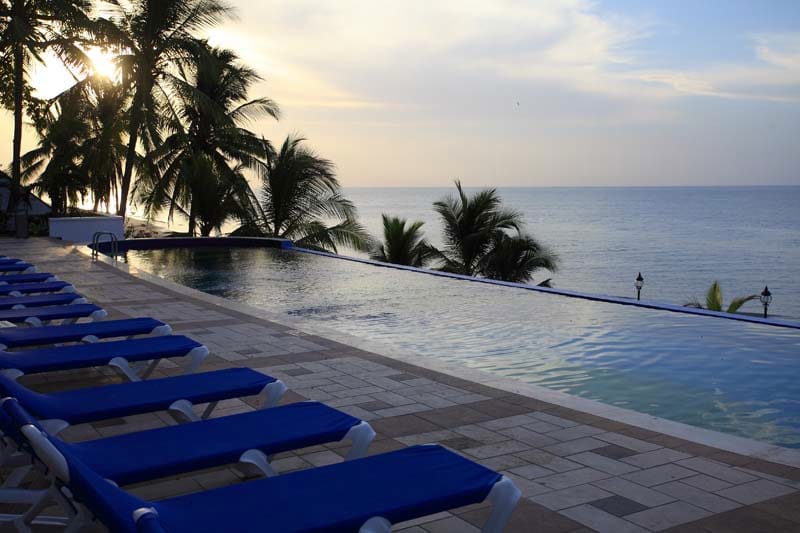 An infinity pool extends towards the horizon, reflecting the colors of the sunset. Sun loungers with blue cushions are positioned along the pool's edge. Palm trees sway gently in the breeze, framing a serene view of the ocean.